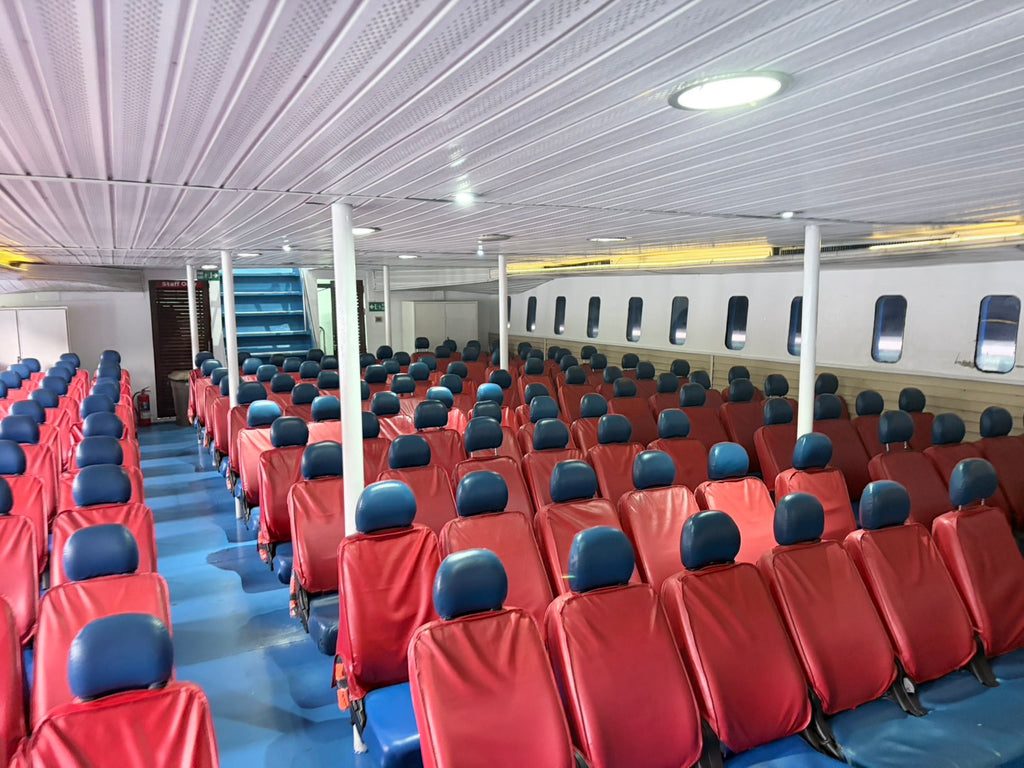 A large interior view of a ship or ferry showing rows of red passenger seats with blue headrests and blue flooring under a ribbed white ceiling.