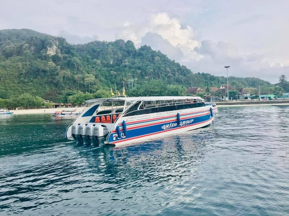 A white passenger ferry with blue and red stripes and the text 'Bali Ferry Group' on its side, sailing on blue water with a lush green mountain and cloudy sky in the background.