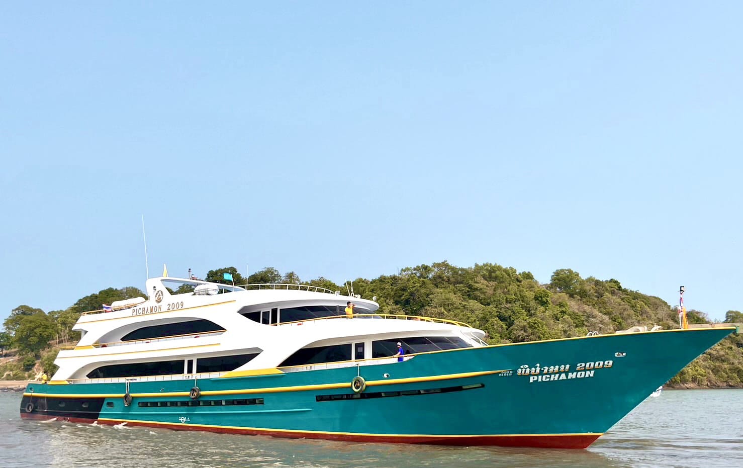 A luxury yacht named 'PICHAMON' in Thai script, painted in teal green and white with gold trim, sailing on calm water with a green forested island in the background under a clear blue sky.
