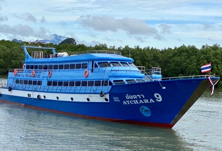 Blue Chukit passenger ferry on the water near a tree-lined shore