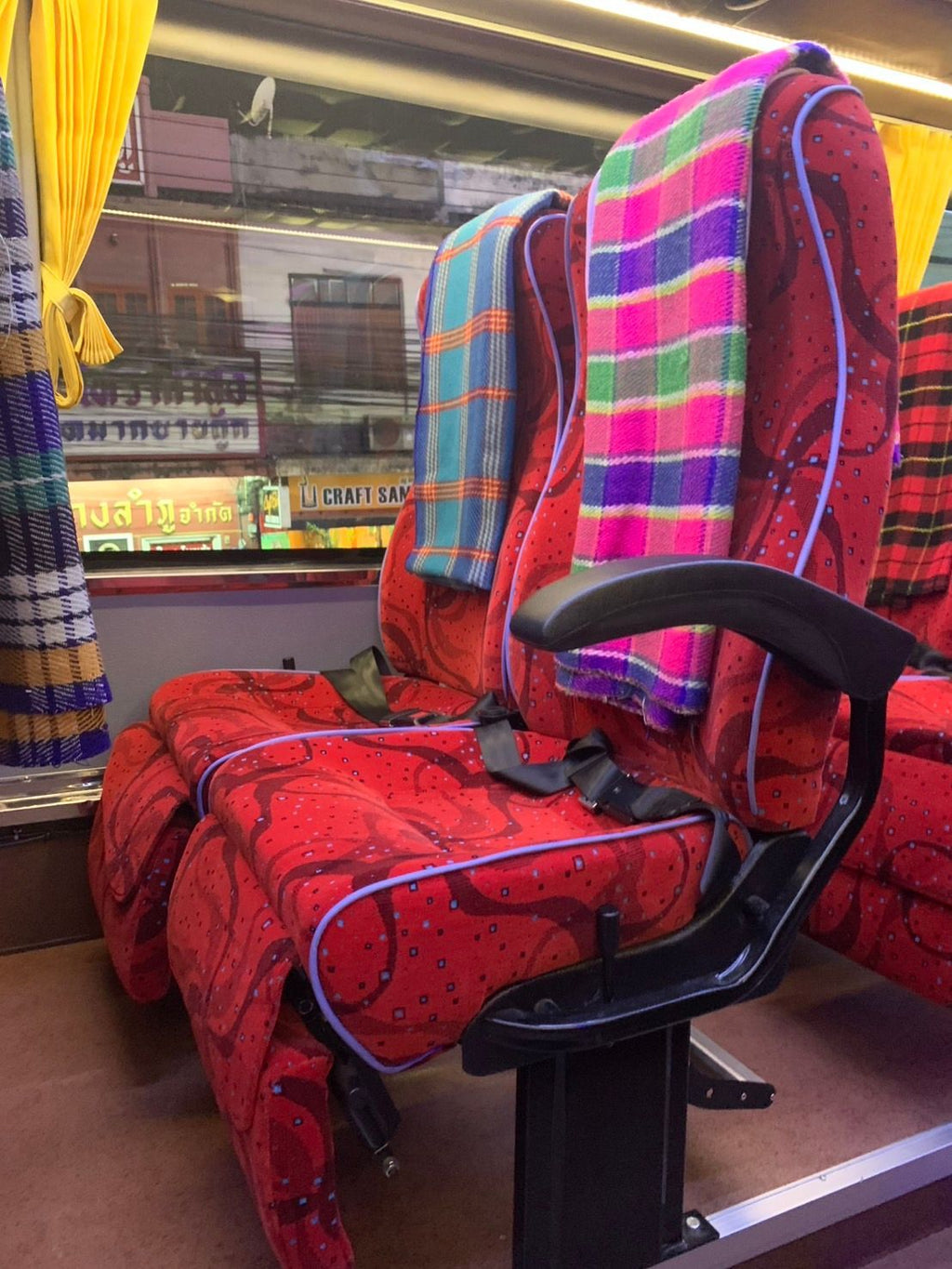 A close-up of a vintage travel seat with red patterned upholstery, a black armrest, and colorful plaid blankets draped over the backrests, set inside a train carriage.