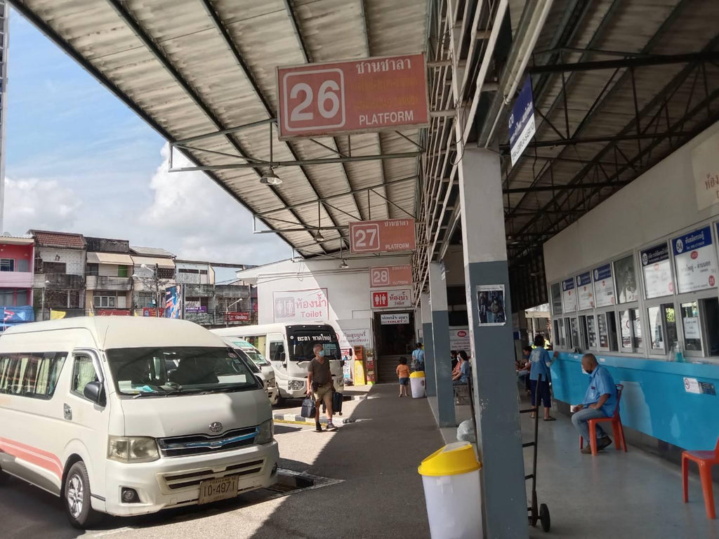 A real-world bus station platform scene with a white minivan parked at platform 26 under a large metal canopy. Signs reading '26 PLATFORM' and '27 PLATFORM' hang overhead, and several people are visible standing or sitting near the ticket windows.