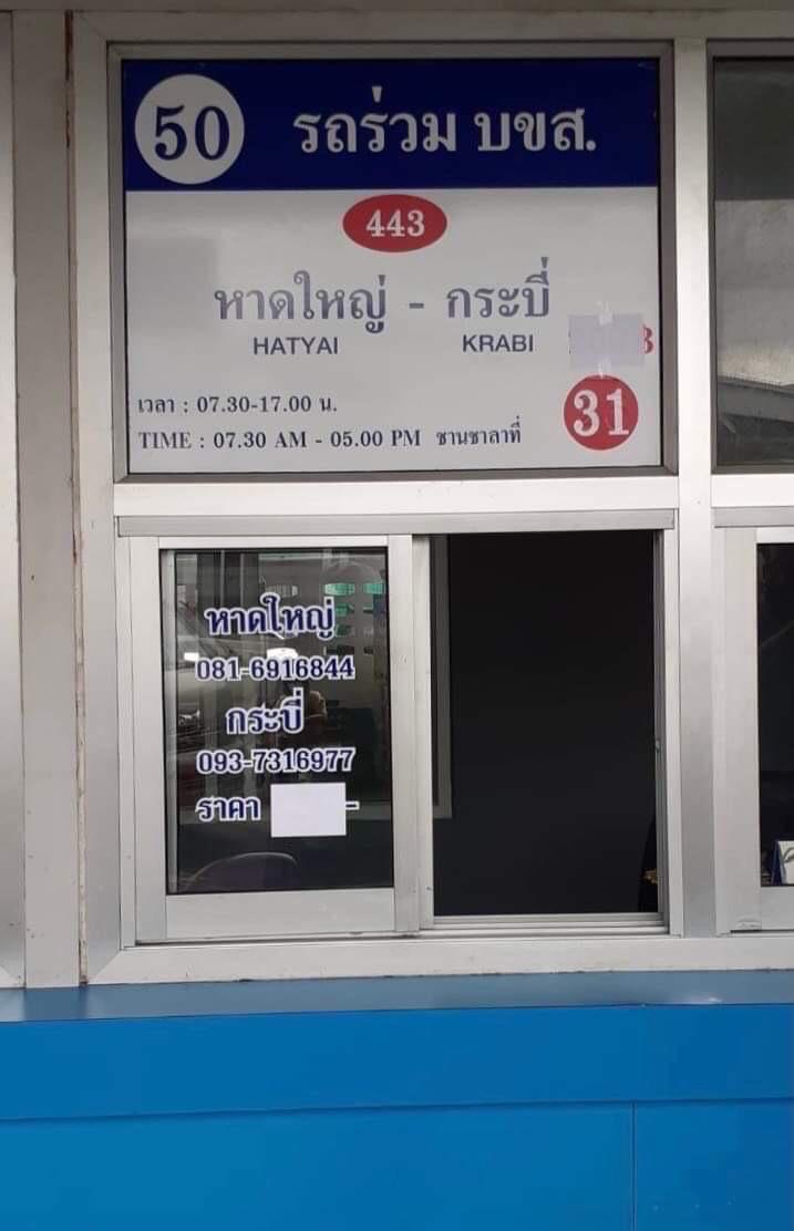 A bus stop sign in Thai with the number 50 and destination Hatyai - Krabi, mounted above a glass window and door at a transportation stop.