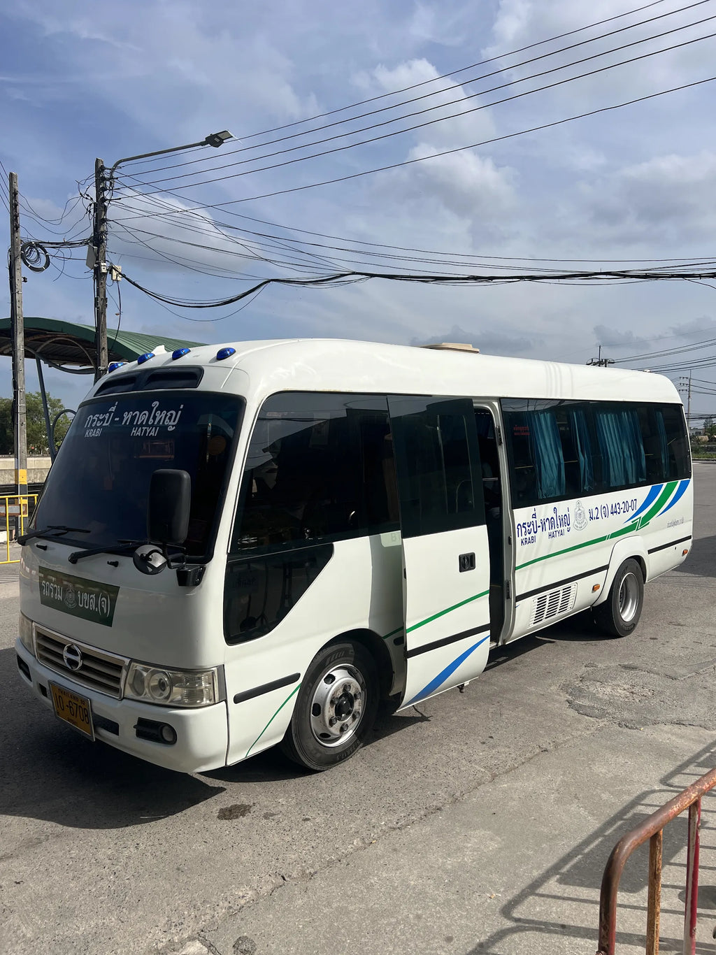 A white minibus with green and blue accents, parked outdoors on a paved area under a cloudy sky. Thai text is visible on the windshield and side door.