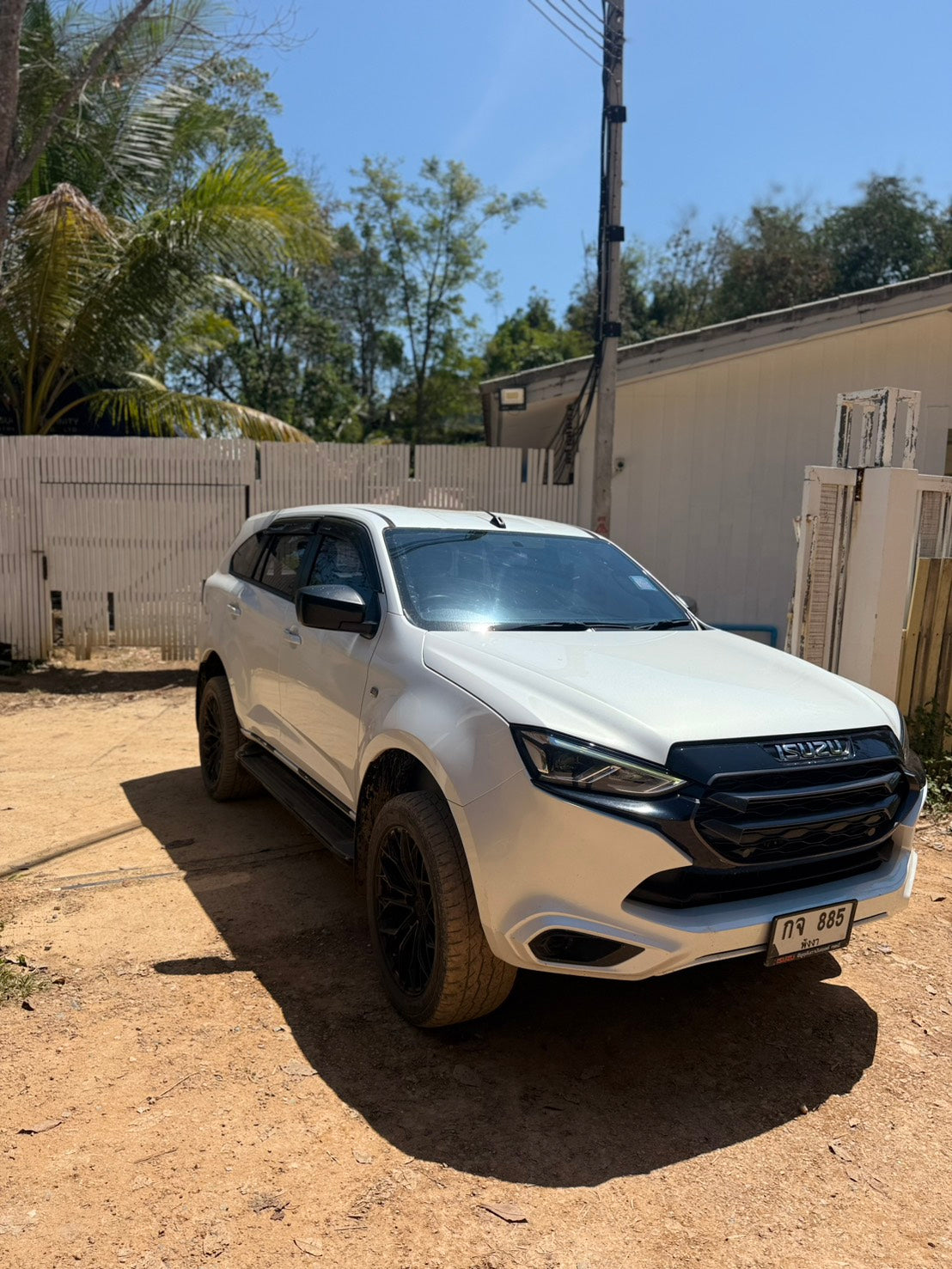 A white Isuzu SUV parked on a dirt driveway in front of a white fence and building under a blue sky.