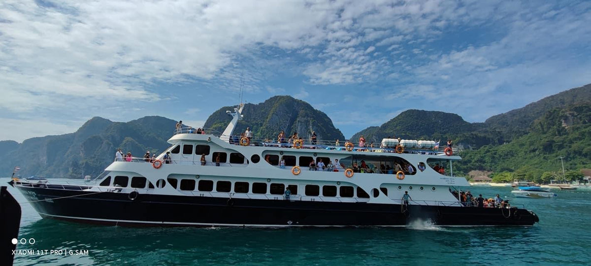 A large white and black passenger ferry or cruise ship sailing on turquoise water with steep green mountains and a blue sky with clouds in the background.