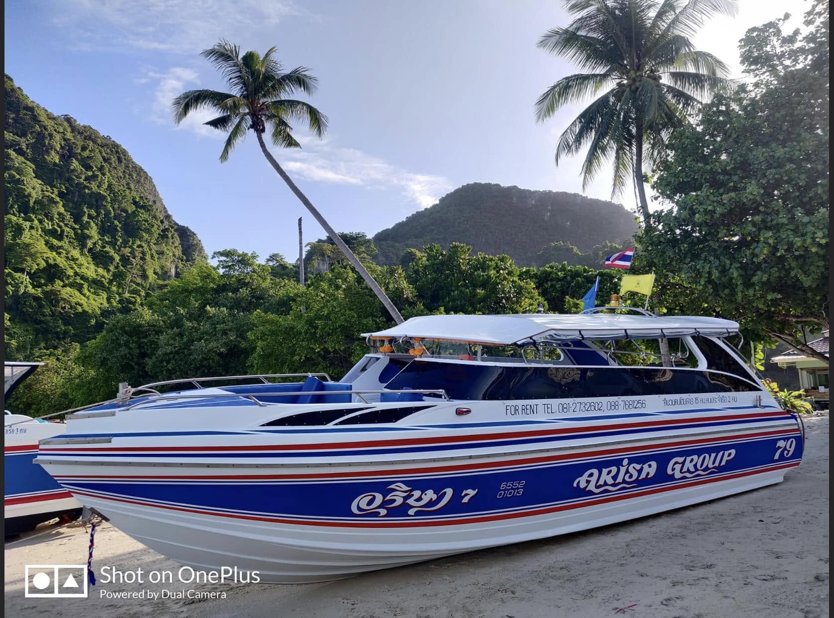 A white speedboat with blue and red stripes and Thai script reading 'Krissan Group' is parked outdoors on a sandy surface with tropical mountains, palm trees, and other boats in the background.