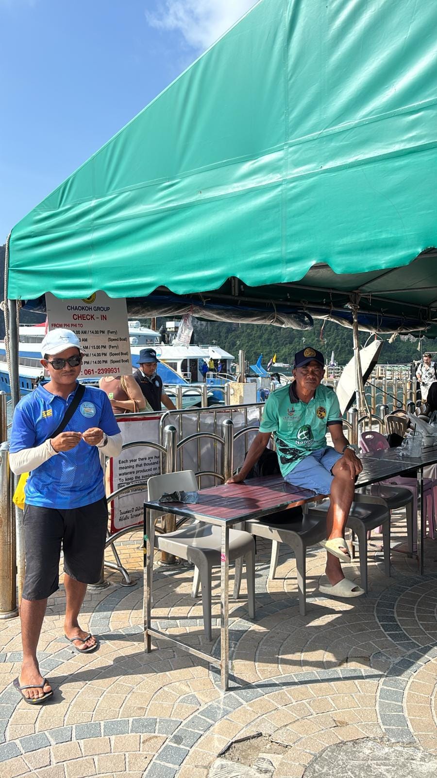 A man in a blue shirt and shorts stands under a green canopy next to a seated man in a green shirt and blue shorts at an outdoor dining area with tables and chairs or Phi Phi Check-in point