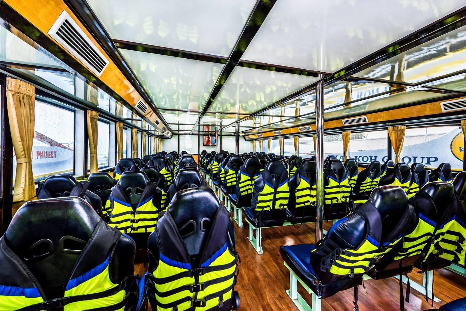 Rows of black and neon yellow life jackets worn by passengers seated inside a large boat cabin with wood flooring and large windows.