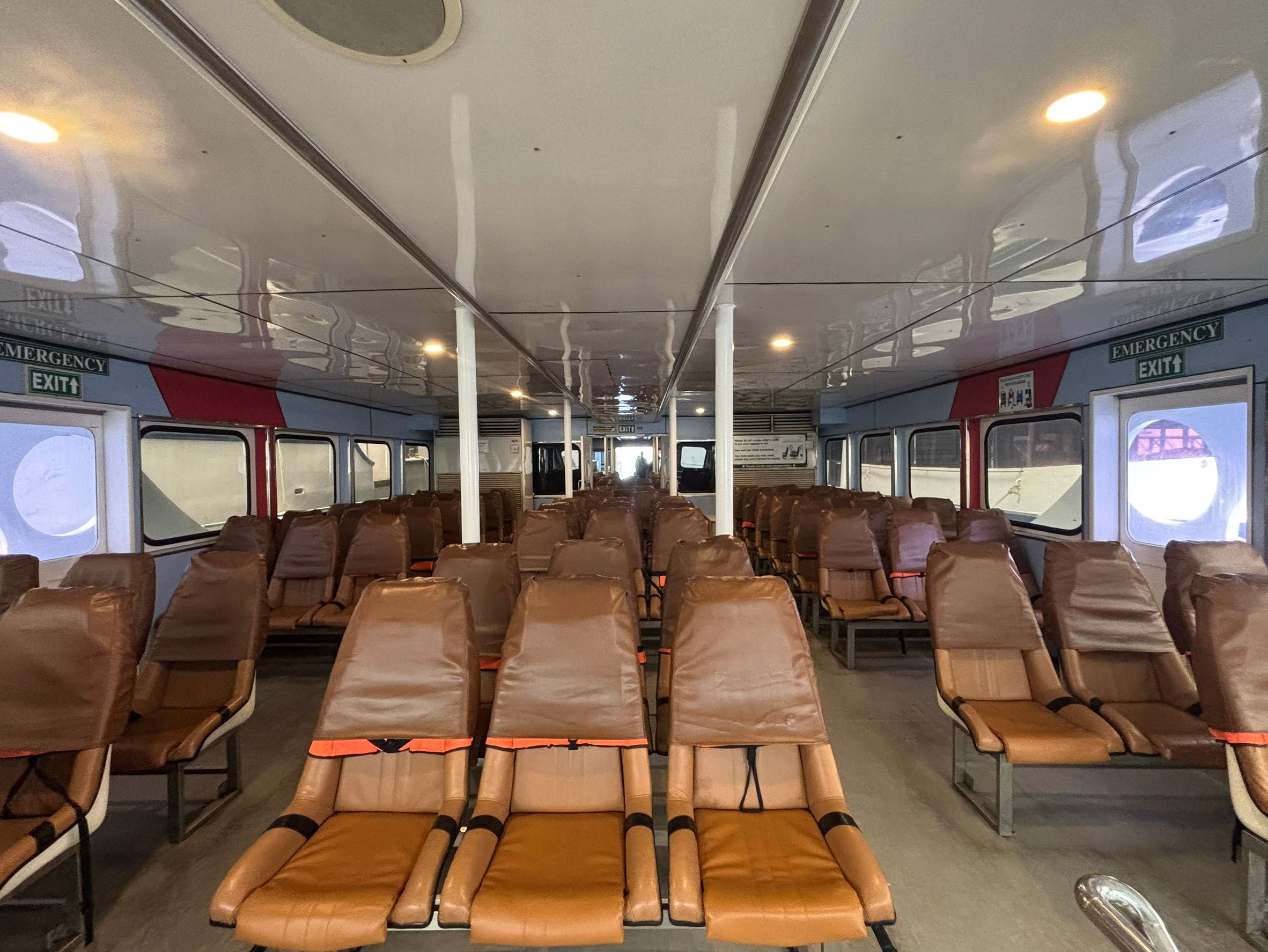 The interior of a train carriage with rows of brown leatherette passenger seats, emergency exit signs, and windows along the side.